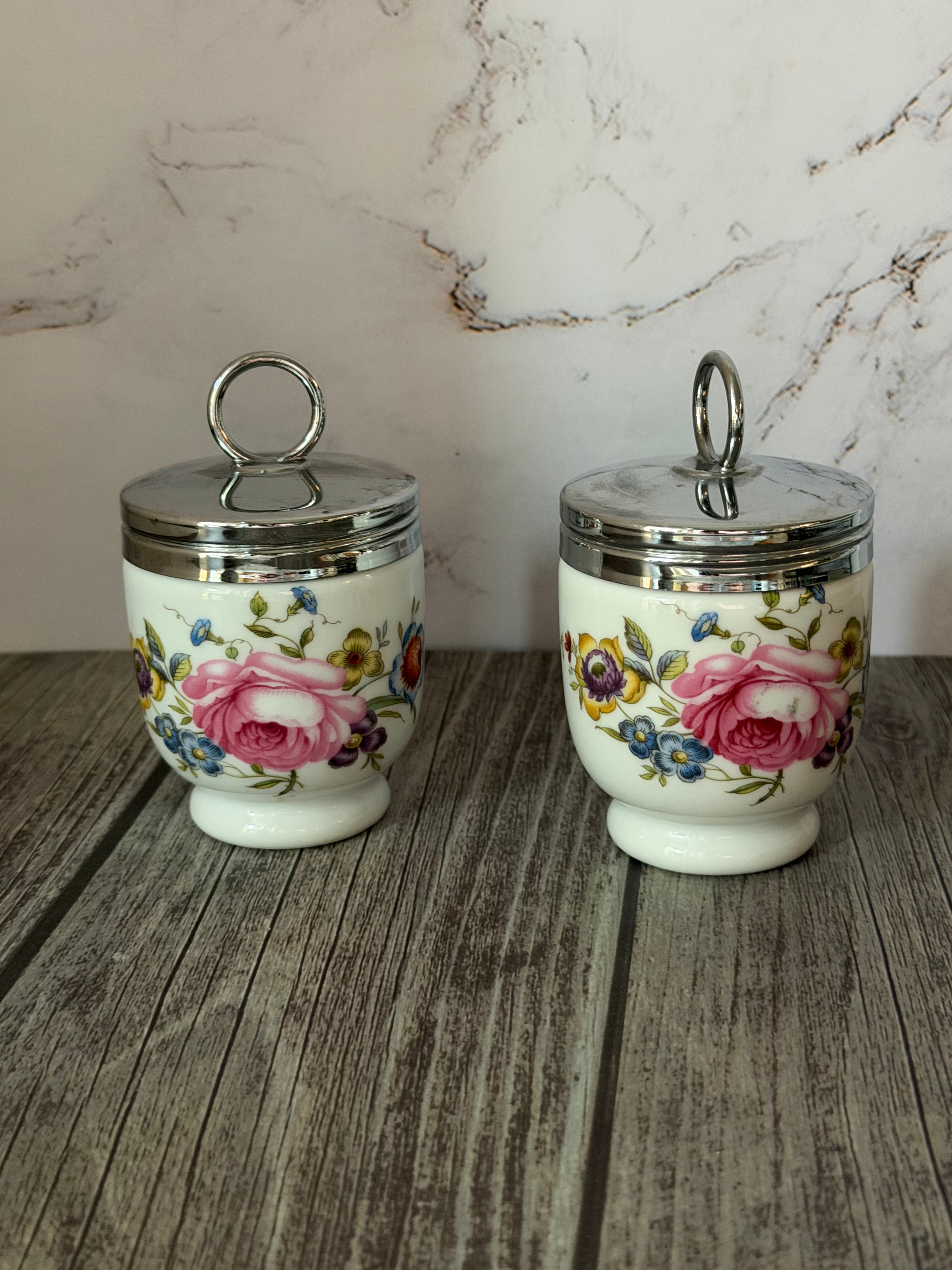 Two floral ceramic jars with silver lids on a wooden surface with a marble wall background.